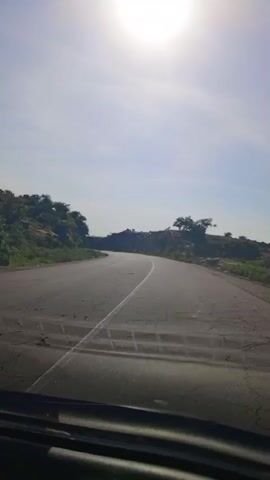 A first-person view from a vehicle traveling on a paved road. The road is curved and appears to be in a rural, mountainous area. The sun is bright and high in the sky, casting a glare. The surrounding landscape is rocky with vegetation and trees. The road has a white line marking the center and shows signs of wear and tear. The vehicle's dashboard is visible in the foreground, reflecting the sky. The time stamp indicates the video is 4 seconds long.