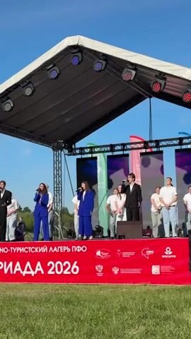 The video shows a group of young people performing on an outdoor stage. Three singers, two women in blue suits and a man in a black suit, stand at the front of the stage with microphones. Behind them are other young people dressed in white t-shirts and light-colored pants who appear to be dancers or backup singers. They are all on a red platform, beneath a stage roof with lighting fixtures. Behind them are LED screens, and banners with Russian text and logos. The text on the red platform reads "ЮНО-ТУРИСТСКИЙ ЛАГЕРЬ ПФО" and "РИАДА 2026". The setting seems to be a sunny day in a park or open field. The group is singing and dancing to the music.