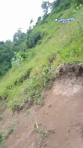 Okay, here's a forensic description of the video:

The video presents a landscape shot of a coastal area.  The camera appears to be positioned on a slightly elevated, grassy, and dirt-covered slope. The view pans to reveal a small waterfall cascading from a rocky cliff face into the ocean. The water is churning around the rocks at the base of the falls. The cliff face is covered in lush green vegetation. Beyond the cliff and waterfall is a hillside that has been cultivated with terrace farms and forest. A few small buildings are visible on the hillside. The sky is overcast.