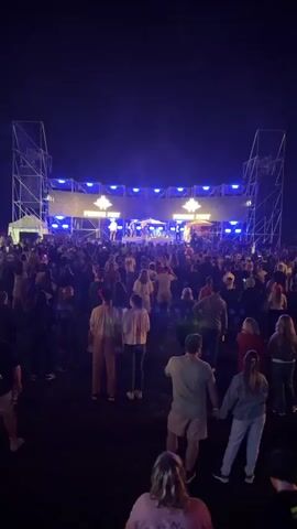 The video shows a nighttime outdoor concert. A large crowd of people are gathered, facing a stage. The stage is illuminated with blue lights and features a band or performers. There are large speakers and scaffolding structures around the stage. The crowd appears to be densely packed in front of the stage. The focus is on the performance and the audience's engagement.
