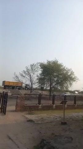 The video shows a historical building, possibly a tomb or mosque, with a dome structure. The building is made of stone or brick, with arches and decorative panels. There are two people in white clothing standing near the building. The surrounding area is a dirt or gravel courtyard.