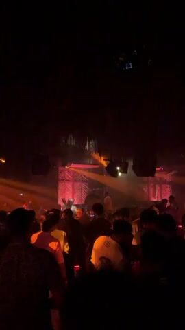 The video shows a dimly lit, crowded indoor space, likely a nightclub or bar. Red and yellow lights illuminate the scene, with beams of light cutting through the hazy atmosphere. Silhouettes of people are visible, some standing and others seated at tables. The focus is on the crowd and the atmospheric lighting, suggesting a lively and energetic environment.