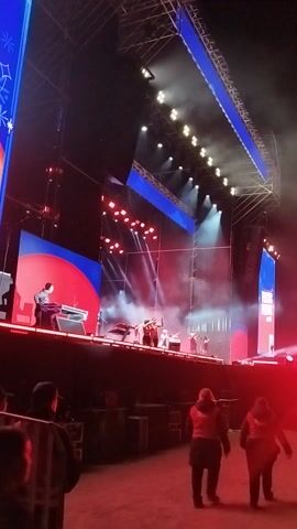 The video captures a vibrant outdoor concert or event taking place at night on a large stage. The stage is framed by metal trusses and features multiple large LED screens displaying dynamic graphics in blue, red, and white. A variety of stage lights, including spotlights, bar lights, and smaller red fixtures, illuminate the performance area, creating an energetic and dramatic atmosphere. A smoke machine generates atmospheric fog, enhancing the visual spectacle.

On stage, several individuals are actively performing. One person is clearly visible playing an electric guitar, while another appears to be operating a keyboard or DJ setup. The performers are brightly lit by the stage lighting.

To the right side of the stage, several people are gathered, likely audience members or event staff, observing the performance. One individual in the foreground has "MAIPÚ" printed on the back of their clothing, indicating the event's location is Maipú, Chile. The scene conveys a lively public gathering centered around a musical performance.