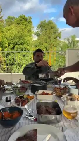Here is a description of the video:
The scene captures a casual outdoor dining setting with multiple people gathered around a table laden with various dishes. A man in a green jacket sits at the table, focused on his plate as he receives a portion of food from another man standing over him. The server is using a utensil to transfer the food from a rectangular metallic container to the diner's plate. The table is covered with a striped tablecloth and holds an assortment of dishes, including salads, roasted vegetables, a pot, a bowl of orange-colored food, and a container of what appears to be meat. A glass of beer stands near the edge of the table. The background reveals a balcony with a metal railing, lush green trees, and a partially visible blue sky with clouds. The atmosphere appears relaxed and social, suggesting a shared meal among friends or family.