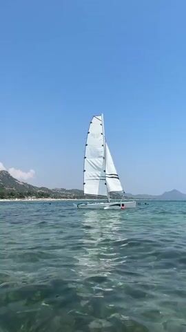 A small sailboat with a white sail is in the water. A person is standing on the boat. There are other boats and people in the water in the distance, along with a beach and hills. The water is clear and the sky is blue.