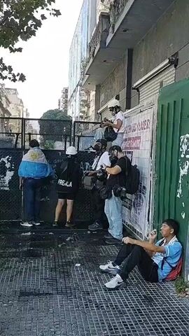 The video depicts a street scene in what appears to be an urban area, possibly during a demonstration or protest. In the foreground, a person is sitting on the sidewalk, wearing a blue shirt, dark pants, and white sneakers, and appears to be smoking. Behind the seated individual, there are several people standing near a black metal barrier. One person has an Argentinian flag draped over their shoulders. Several others are wearing helmets and gas masks and appear to be either press or part of the protest. One person is wearing a jacket with "PRENSA" written on the back. Some individuals are holding cameras. A building is visible behind the barrier, with a sign that says "POLICIA DE LA CIUDAD," indicating it is likely a police station. The sky is bright, and a tree with green leaves is visible in the upper left of the frame. Overall, the scene has a tense and uncertain atmosphere.