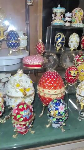 The video pans across a display case filled with ornate, jeweled decorative items. The items are primarily Fabergé-style eggs, miniature carousel horses, Ferris wheels, crowns, and various animal figurines, including cats and frogs. Many of the pieces are gold-toned with colorful enamel and faux gems. The display is lit and set against a dark background, with shelves made of glass and wood.