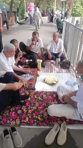 Six men are sitting on blankets on the ground, eating and talking in an outdoor setting. They are wearing traditional Muslim head coverings. Some men are eating fruit from a platter. A thermos and other items are spread out on the blankets. In the background, other people are walking by and some are holding balloons. The scene appears to be a park or recreational area.