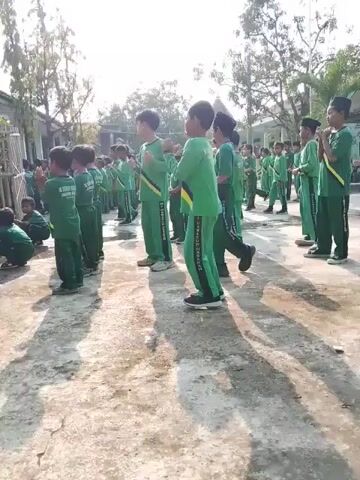 A large group of children, all wearing green uniforms, are standing in rows in an outdoor area. Some of the children are moving their arms or legs, while others appear to be standing still. The area they are in seems to be a courtyard or similar open space, with trees and buildings visible in the background. The weather appears sunny. Some of the children are wearing sashes.