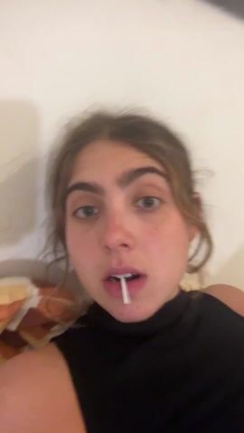 A young woman with fair skin, light brown hair pulled back, and a black sleeveless turtleneck is centered in the frame. She has a lollipop in her mouth. She removes the lollipop, speaks to the camera, and pauses while looking around. The video is shot at a close-up angle, and the woman appears to be lying down with a pillow visible behind her head and shoulder. The background is a plain white wall, and some part of the ceiling can be seen in the upper right corner of the video.