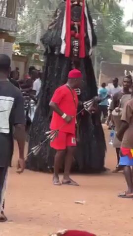The video shows a tall, black, shrouded figure standing in a sandy area. It appears to be some sort of ceremonial costume. The figure has a face covering with white and red decorations and what look like small masks attached. A person wearing a red outfit and cap walks in front of the figure, holding a spear or staff. People surround the figure, and a man in the background is carrying a drum. Some of the people are looking at the figure while others are just standing nearby. Buildings and trees are visible in the background.