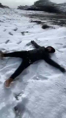 The video shows a person lying on their back in a snow-covered landscape, making a snow angel. The person is wearing dark clothing, including a jacket, pants, gloves, and a hat. They are also wearing brown boots. The person is lying with their arms and legs outstretched. The ground is covered in white snow with footprints. In the background, there are some dark objects that appear to be logs. The sky is overcast. The person appears to be smiling. The video appears to have been taken from a slightly elevated angle.
