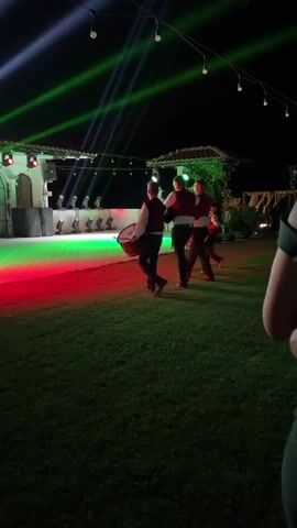At night, four young males are seen performing a traditional dance with a drum on a grass field. They are wearing matching outfits, consisting of vests and shirts, and appear to be moving in unison. The background features a stage illuminated by colorful spotlights that cast beams of light into the night sky. There are also string lights and decorative elements that suggest an outdoor event or festival. The stage itself has a large screen and what appear to be speakers, indicating a musical performance. The lighting is dynamic, with red and green lights illuminating the performance area, creating a vibrant and festive atmosphere.