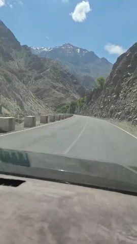 A static shot from inside a car shows a straight, paved road running through a mountain pass. Concrete barriers line the road on both sides. The mountains are rocky and sparsely vegetated. In the distance, a snow-capped mountain can be seen under a partly cloudy, blue sky. Music is playing.
