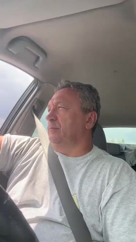A man in a grey t-shirt is driving a car. He is looking to the left and then up. The sun is shining.