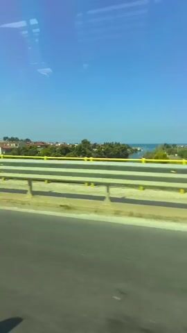 The video shows a view from a vehicle moving along a road. A concrete barrier with yellow markings separates the road from a landscape beyond. This landscape includes houses, trees, and a body of water, likely the sea, visible in the distance. The sky is clear and blue, indicating a sunny day. The movement suggests the camera is positioned inside a moving vehicle, possibly a bus or a car, looking out the window.