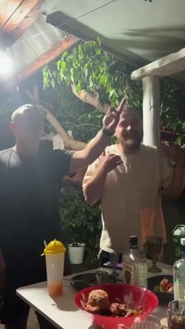 Two men are standing at a table outdoors under a covered structure. The man on the left is wearing a dark t-shirt and shorts, and the man on the right is wearing a light-colored t-shirt and appears to have a beard. Both men are smiling and gesturing. On the table, there are bottles of alcohol, glasses, and food. Other people are partially visible in the foreground. The environment appears to be a backyard or patio.