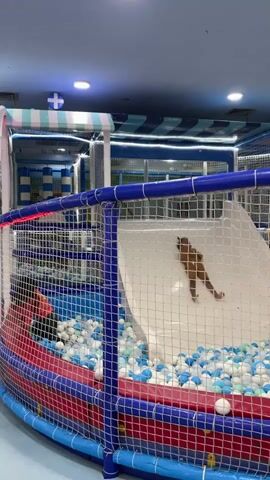 A child slides down a white plastic slide into a ball pit. The ball pit is filled with blue, white, and light blue plastic balls, and is contained within a structure resembling a boat. The boat structure has red and blue netting. The environment appears to be an indoor playground.