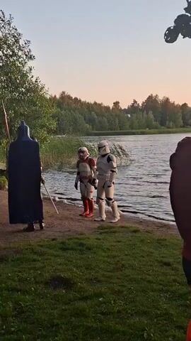 The video shows a group of people by a body of water at dusk. Several individuals are dressed in costumes resembling characters from Star Wars. One person is dressed as Darth Vader, complete with a cape and helmet, and is holding a lightsaber that appears to be red. Another person is in a Stormtrooper costume. There are also individuals dressed as Rey and another Stormtrooper, along with a few people in casual clothing. The scene appears to be a gathering or a reenactment, possibly a Star Wars fan event. The environment is natural, with trees and grass surrounding the water. The lighting suggests the sun is setting.