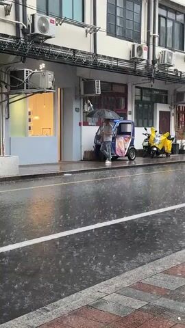 A person walks down a wet street holding an umbrella. It is raining heavily, and the street is covered in puddles. A blue three-wheeled vehicle is parked on the sidewalk, and a yellow scooter is parked behind it. The buildings in the background have multiple stories and windows. The scene appears to be in an urban environment.