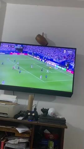 A television screen displays a soccer match. The game is in progress with players on the field. The score is visible on the screen, along with team names. The crowd in the stadium is visible in the background. The television is mounted on a wall above a shelf unit. On the shelf unit, there are various objects including a printer, a game controller, and other electronic devices. A decorative object is also hanging on the wall above the television.