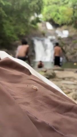 A video shows a caterpillar on brown fabric, with a blurred waterfall and three people in the background. The caterpillar is small, fuzzy, and light brown. The fabric is wrinkled. The waterfall is a series of cascades falling into a pool. The people are shirtless and wearing shorts or swim trunks, and appear to be swimming or wading. The background consists of trees and rocks.