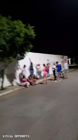 At night, a group of about a dozen people stand on the sidewalk next to a street, in front of a long white wall with painted images. Some are sitting on the curb.  A tall man is standing and painting something on the wall with a brush or stick.  A few other people are watching or talking. The image is a bit blurry and not very high definition. There is a tree to the left.  The sky is black, and there is a street light in the distance to the right.  The camera is still.