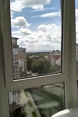 The video shows a view from a high-rise apartment window overlooking a neighborhood. A soccer field is visible below, and other apartment buildings and trees are in the distance. The sky is blue with some clouds. A white round table is near the window. A cat is lying on a gray stool, cleaning its paw.