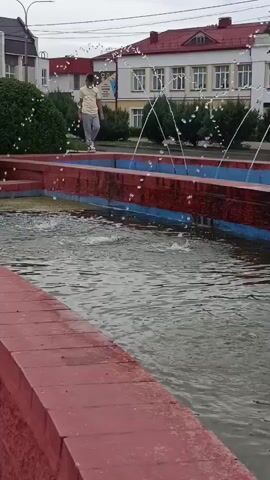 A person on roller skates is standing at the edge of a fountain. The fountain has multiple jets of water spraying upwards. The person is wearing a light-colored shirt and dark pants. In the background, there is a building with red roofs and white walls, as well as trees and a street with cars. The sky is overcast.