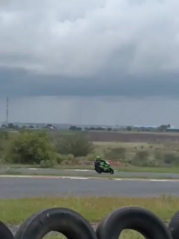 The video shows two motorcycles racing on a track. In the foreground, there are three black tires. The first motorcycle is predominantly green and white, while the second is orange. The race is taking place on a paved track, with a grassy border. Beyond the track, there is an open field with sparse vegetation and buildings in the far distance under a gray, cloudy sky.