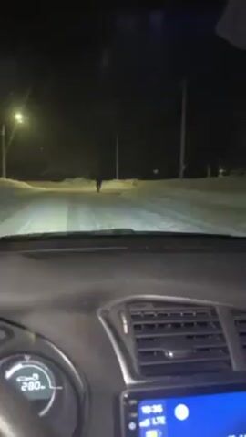 A video shows a nighttime view from inside a car driving on a snowy road. A person is walking on the road ahead of the car. As the car approaches, the person steps off the road and stands on a snow bank to the side. The person is wearing dark clothing and appears to be wearing skis or snowshoes. The car continues past the person. The dashboard of the car is visible, with the radio playing "Beautiful Pain" by Eminem and Sia.