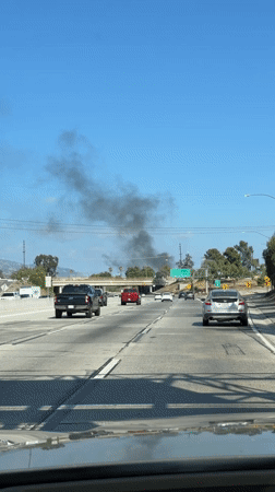 Dark smoke rises along a Los Angeles highway under clear skies.