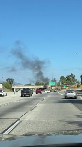 Dark smoke rises along a Los Angeles highway under clear skies.