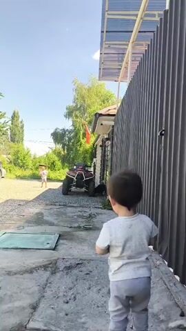A boy in a grey shirt and pants stands with his back to the camera, facing a grey metal fence. To the left, a child in a white shirt and shorts runs on a gravel path. Behind the running child is a red ATV. Trees and a building are visible in the background under a bright blue sky.