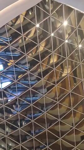 The video shows a geometric ceiling pattern made of triangular reflective tiles with white trim. The reflections in the tiles show interior scenes with bright lights and screens, giving a distorted view of the space below. The camera angle is pointed upwards towards the ceiling.