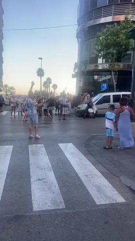 A group of people, some wearing Argentina national football team jerseys, are gathered on a street corner near a pedestrian crossing. Two individuals are playing drums in the middle of the street, one with a larger drum and the other with a smaller one. A silver van is parked on the side of the road, and several people are standing near it, watching the drummers. Other people are visible in the background, some with their arms raised. Palm trees are visible behind the crowd. The sky is clear and appears to be dusk.
