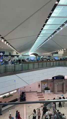 The video pans across a bright, modern airport terminal. The architecture features a high, vaulted ceiling with integrated lighting. The perspective is from an upper level looking down and across. On the upper level, a row of information or check-in desks is visible, staffed by several individuals who appear to be employees. Travelers are interacting with these staff members. Below, on the main concourse, a group of people are waiting, some seated and others standing. To the left, on the lower level, there is a sign that reads "INFORMATION CENTER." There are also decorative elements and people in cultural attire on the lower level, possibly performers. The overall environment is clean, spacious, and functional, typical of a contemporary airport.