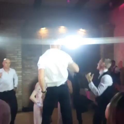 The video shows a group of people dancing in a dimly lit room. There are several bright lights illuminating the scene, creating a hazy atmosphere. In the foreground, a man in a white shirt and black pants is facing away from the camera, appearing to dance. To his left, another man in a white shirt and black pants is also dancing and pointing his finger upwards. To the right of the man in the center, a man in a white shirt and black vest is dancing with a woman. Further back, more people are visible dancing and socializing. The walls appear to be made of brick, and there are lights mounted on the ceiling. The overall impression is of a lively party or reception.