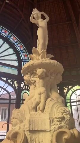 The video opens with a close-up of a large, ornate fountain statue in an indoor setting. The statue depicts a female figure at the top, arms raised, and several cherubic figures below her. The fountain is made of a light-colored stone or plaster. Behind the fountain, a large, arched stained-glass window is visible, featuring a circular design with red and blue accents. The ceiling is made of dark wood with visible beams. The camera then pans upwards and to the right, showing more of the wooden ceiling structure and the stained-glass window. The view shifts to focus on the architectural details of the space, including the wooden arches and beams, and more arched windows, some with green-tinted glass. The camera continues to pan, revealing more of the interior space. The last shot shows a large mural on the wall to the left, depicting a landscape scene. The overall environment appears to be a grand, old building, possibly a pavilion or public space. There are no visible people or discernible actions in the video.