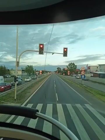 This video captures a view from a vehicle, likely a bus or a large truck, looking forward through the windshield. The perspective suggests the vehicle is stopped at a traffic light, indicated by the red signals visible in both the foreground and background. In the immediate foreground, a portion of the steering wheel and dashboard is visible. The road ahead is a multi-lane highway, with a pedestrian crossing marked by white stripes directly in front of the vehicle. Cars are visible in the distance, also appearing to be stopped. On the right side of the road, there's a large billboard advertising "Jurajska" bottled water, depicting a volleyball player. A traffic light and various road signs are also present near the billboard. The sky is overcast, suggesting a cloudy day. On the left side of the road, there are trees, parked cars, and some construction activity with machinery visible. The video appears to be a continuous shot without any significant edits or camera movements beyond what would naturally occur in a vehicle at a standstill.