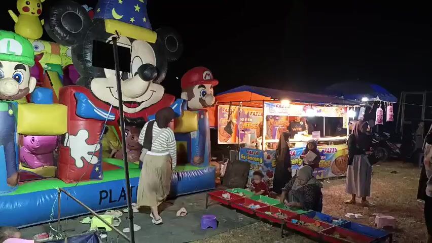 The video shows an outdoor night scene with a large inflatable bouncy castle featuring characters such as Mickey Mouse, Luigi, and Mario. Several people are present, including adults and children. Some individuals are standing near the bouncy castle, while others are gathered around food stalls. One stall has colorful signs advertising food items. There are also some game stalls with rows of colorful prizes. The ground appears to be dirt or grass. The lighting is dim, with artificial lights illuminating the stalls and bouncy castle. A few vehicles are visible in the background.