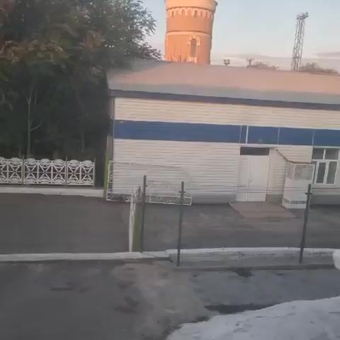 The video shows a street scene with a building in the background. The building is white with a blue stripe and has a white door. There is a fence in front of the building, and a gate. A water tower and a radio tower are visible in the distance. The sun is setting, casting a warm glow on the scene. The video is shaky, suggesting it was filmed handheld.