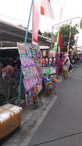 A street vendor displays a variety of colorful toys on a stand. Several flags, including the Indonesian flag, are flying in the background. People, some wearing headscarves, are seen browsing the merchandise. A box wrapped in foil is in the foreground.