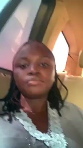 A woman is filming a selfie video from inside a vehicle. She has dark skin, a nose ring, and dreadlocks. She is wearing a gray top with white lace detailing around the neckline. The interior of the car is beige. The window and a glimpse of the outside world can be seen in the upper left corner of the frame. The lighting appears to be natural daylight. The woman looks directly at the camera with a slightly serious expression.