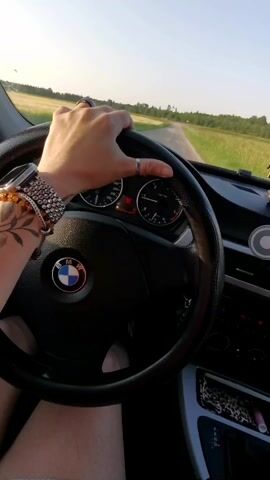 The video shows a first-person perspective of someone driving a BMW. The driver's hands are on the steering wheel, with a tattoo visible on the left arm. The dashboard displays include the speedometer and other indicators. The car is on a dirt road, surrounded by fields of grass and trees under a clear sky. The sun appears to be setting, casting a warm glow on the scene. The overall impression is one of a leisurely drive through a rural landscape.