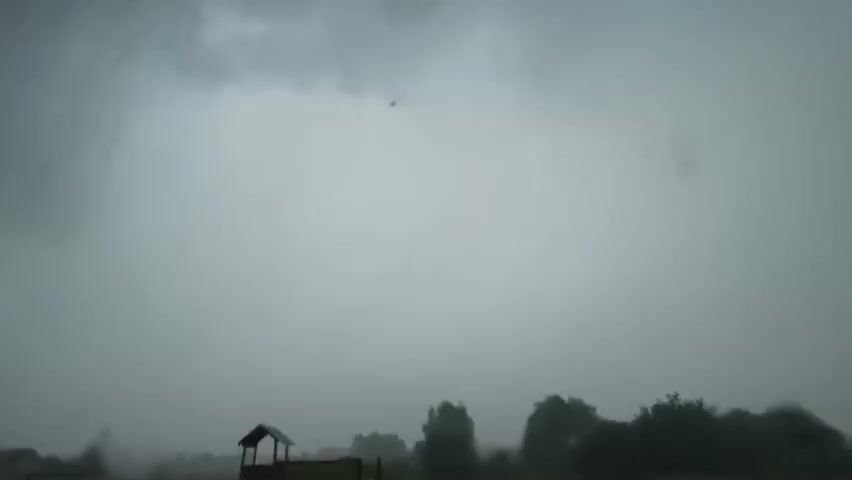 This is a video of a thunderstorm. The scene is set in a rural location in Kyviškės, Lithuania, with a treeline and a small structure resembling a playground or a shed in the foreground. The sky is overcast with dark clouds, indicating a stormy atmosphere. The weather is characterized by heavy rain and lightning, as evidenced by a bright flash illuminating the sky with a purplish hue during one segment. The overall mood is dramatic and somewhat ominous due to the intense weather. No people are visible in the footage, and there is no visible text. The time of day appears to be either late afternoon or evening, given the dim lighting conditions before the lightning strike.