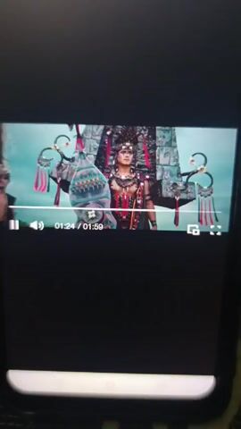 Alright, here's a description of the video:  The video shows a black cellphone screen. On the phone's screen is a video. The video appears to be a music video on the phone screen. The video shows three people, the person in the middle appears to be female with two men on either side of her. All three are wearing ceremonial style clothing and headwear. In the background are objects that look like temple decorations.