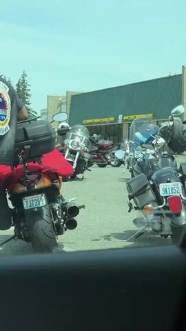 The video shows a group of people standing around motorcycles in a parking lot on a bright, possibly sunny day. Two individuals in the foreground are wearing black leather vests with patches that say "CHAPTER ONE AMERICAN VETERANS AMVETS DEPT OF WA". The person on the left is wearing a blue baseball cap, a grey t-shirt, and dark pants. They are holding a large grey water bottle. The person on the right is wearing a bandana with a red and white pattern, a green long-sleeved shirt, and blue jeans. Behind them, several motorcycles are parked, including a dark red one in the middle foreground with a license plate that reads "4US567". In the background, there are more motorcycles and a building with a green facade and yellow accents. The environment appears to be an outdoor gathering, possibly related to a motorcycle club or event for veterans. The camera seems to be positioned inside a vehicle, as evidenced by the dark interior framing the shot.