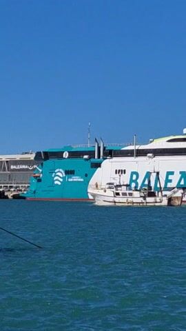The video shows a bright, sunlit view of a harbor scene. In the foreground is the turquoise sea, its surface slightly rippled. A small, white fishing boat is visible, moving across the water in front of a large, docked ferry. The ferry has the word "BALEÀRIA" written on its side in bold, green letters. Part of the ferry is white, while another section is a vibrant turquoise color, featuring a graphic of a wave or water symbol and the words "GAS NATURAL." The sky above is a clear, cloudless blue. A fishing line is visible along the left edge of the frame.