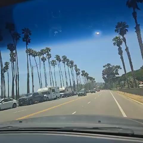 Here is a detailed description of the video:

The video shows a car driving down a coastal road lined with palm trees on a sunny day. The sky is bright blue with a few clouds, and the sun is shining intensely. The road has two lanes and a yellow dividing line. There are parked cars, including several RVs, on the left side of the road, along with moving cars on the road. A few larger trees, in addition to the palms, are visible on the right side. The video is shot from inside the moving car through the windshield. Music is playing, as well.